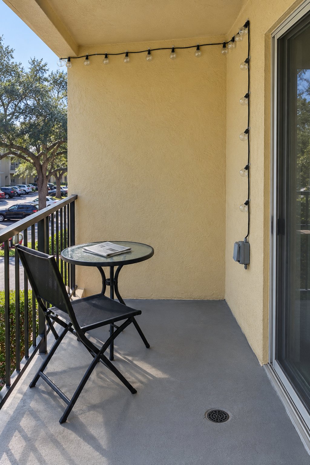 Balcony with an outlet, one strand of outdoor-rated string lights along the back wall at dusk, the cord kept off the floor, one chair and a small table