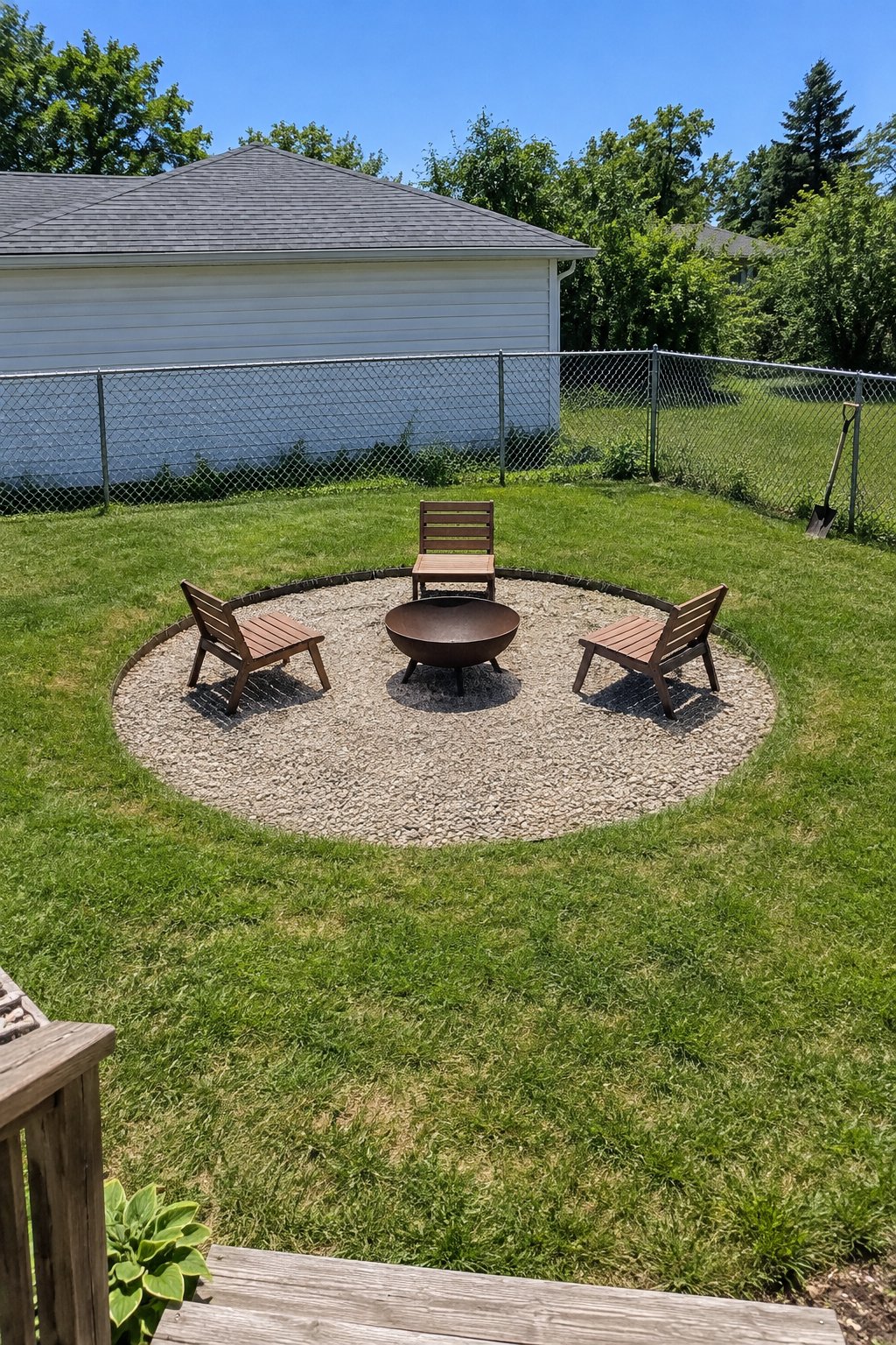 Gravel fire pit area sunk just below lawn level in a backyard corner with steel edging, a steel fire bowl in the center and three low chairs