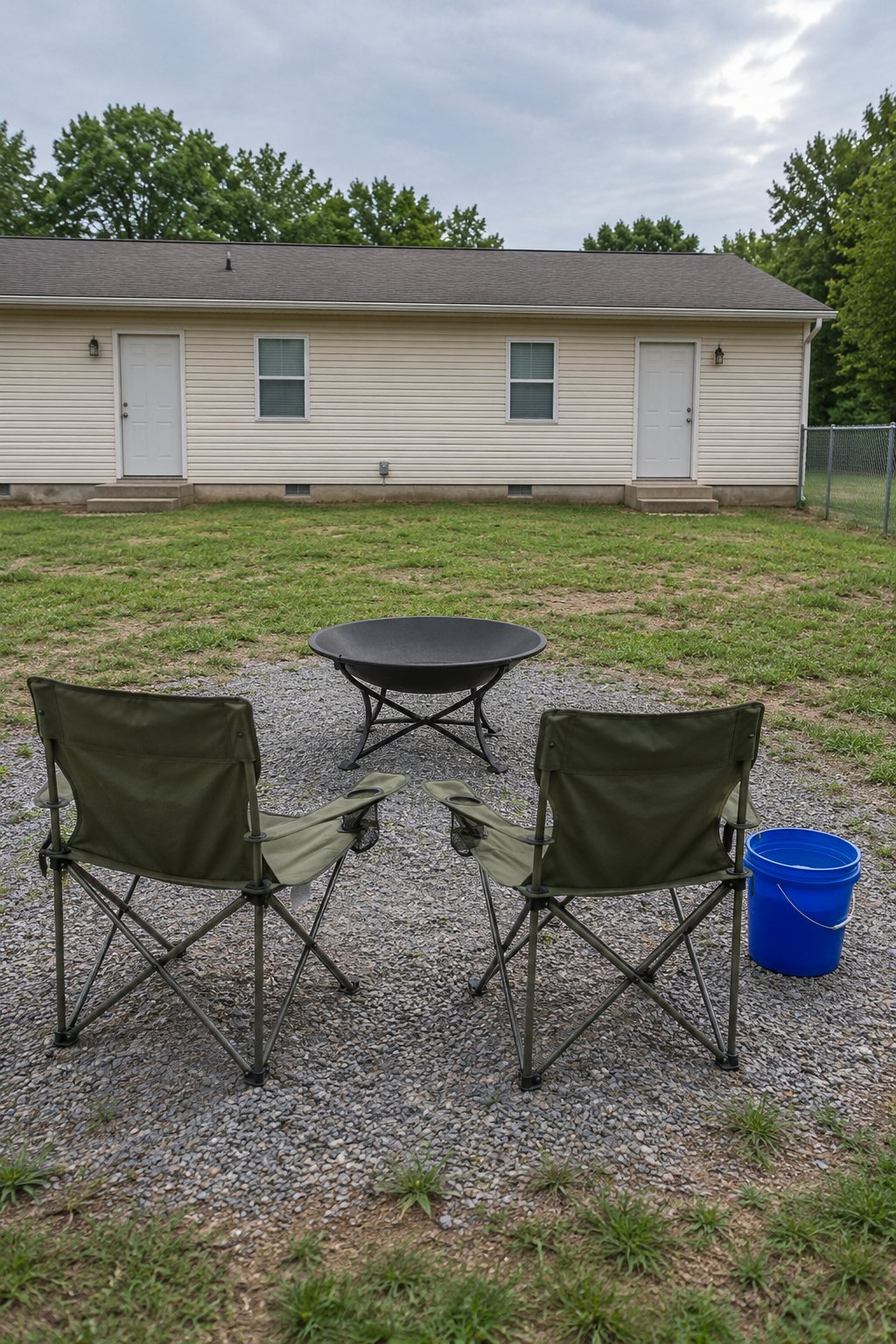 Small firepits backyard ideas for renters, a steel fire bowl on folding legs with two folding camp chairs on a gravel patch behind a rental duplex